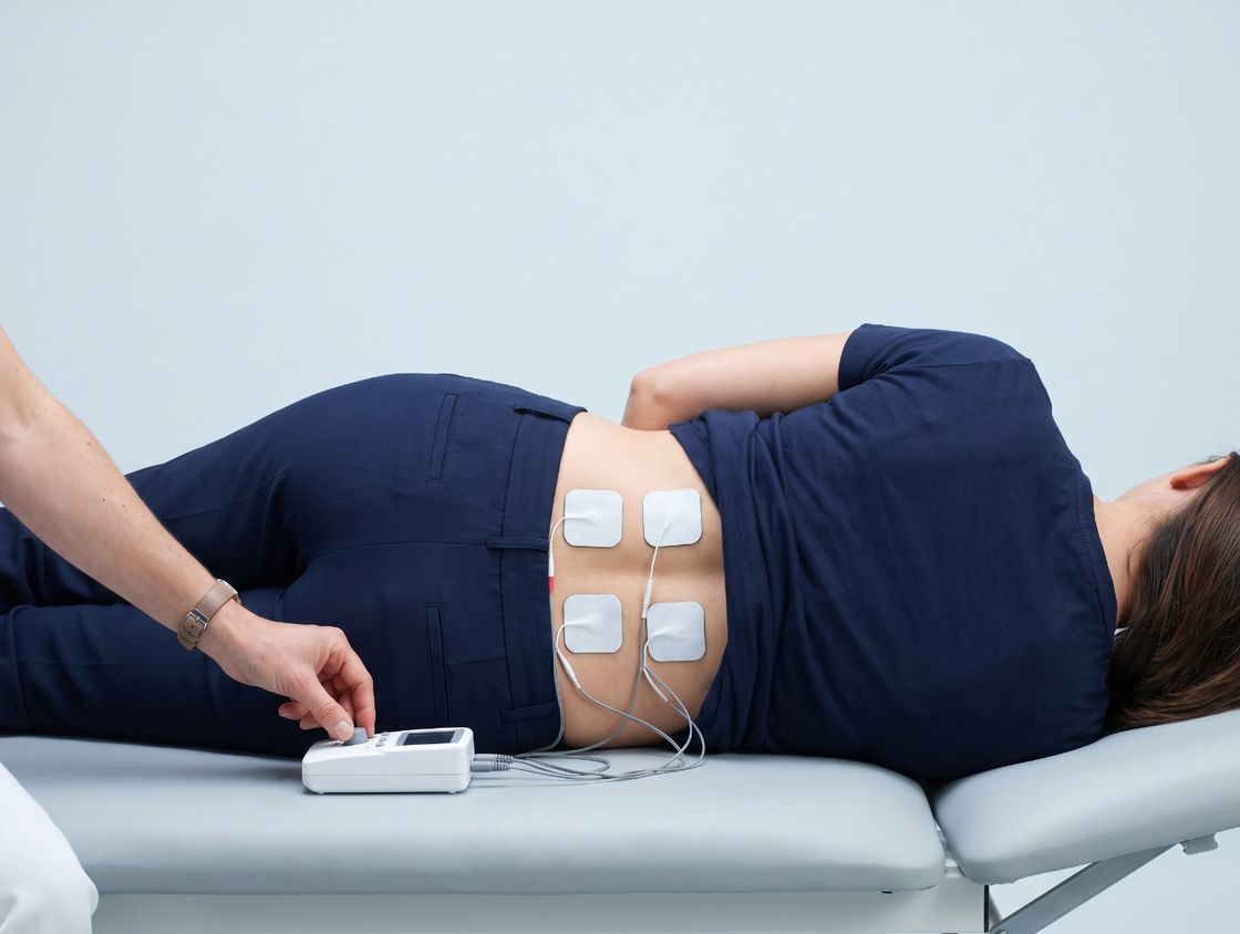 A patient lies on her side with four electrode pads on her lower back while the chiropractor adjusts a small electrotherapy unit on the table.